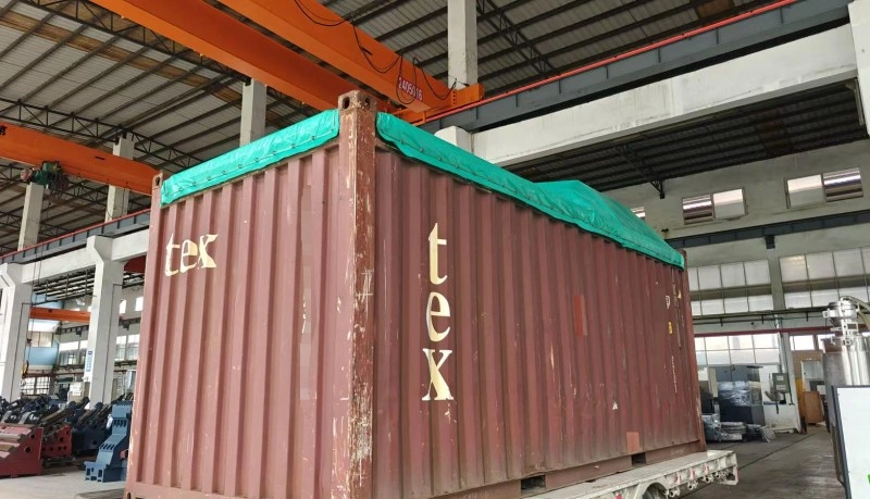 A large industrial machine wrapped in green protective tarp being loaded into a red shipping container inside a factory warehouse.