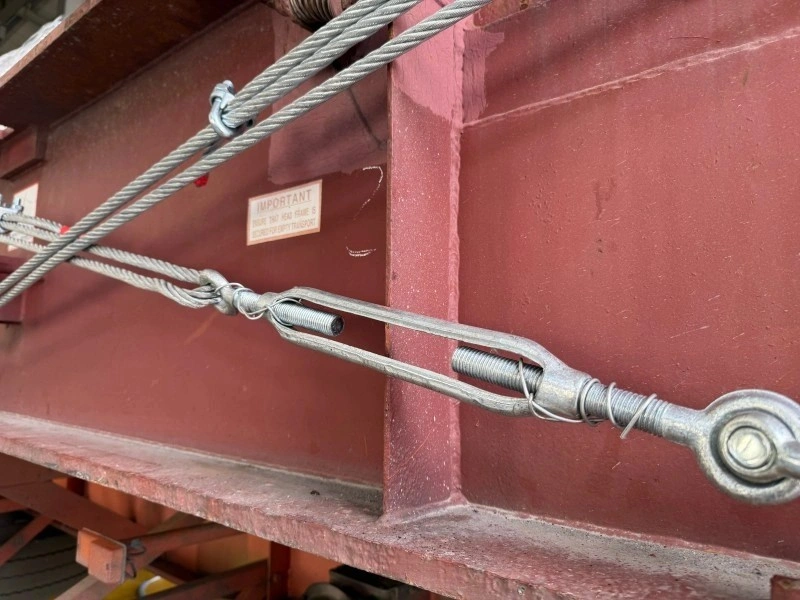 Close-up view of galvanized turnbuckles and wire rope assemblies attached to a red steel container frame, highlighting the precision hardware used in heavy cargo restraint systems for project freight.