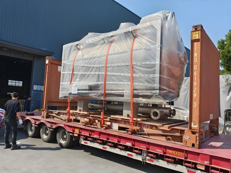 A large machine wrapped in clear industrial barrier film and secured with orange lifting straps sits on a flatbed chassis beside an open-top container frame, ready for sea freight export.