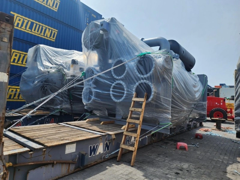 A large industrial water treatment unit, wrapped in protective plastic and secured with steel cables, is loaded onto a blue flat rack container at a port yard, with stacked shipping containers labeled “TAILWIND” in the background.
