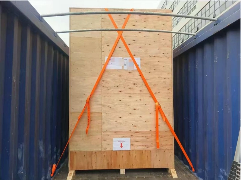 Inside a Wan Hai shipping container, a tall wooden crate is braced with crossed orange tension straps and marked with a downward arrow label for correct orientation during unloading.