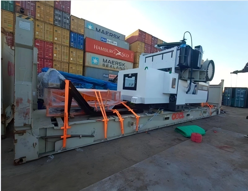 A massive white CNC machine is positioned on a grey flat rack container in a busy port terminal. The machine is secured with multiple orange lashing straps attached to the container's strong points. In the background, stacks of shipping containers from various companies like Maersk and Hamburg Süd are visible under a clear sky.