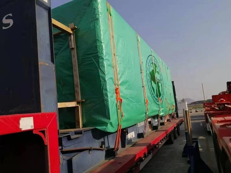 A large, rectangular piece of cargo, wrapped in a green waterproof tarpaulin and reinforced with wooden beams, is securely strapped onto a red flatbed trailer using orange ratchet straps. The scene is set outdoors in a port or industrial area under a clear sky.