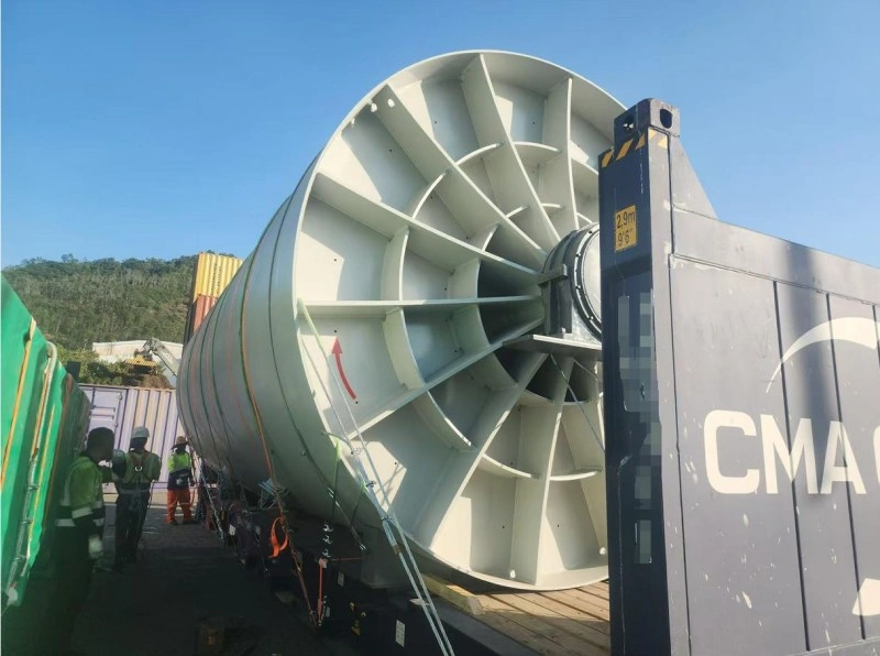 A massive, light-gray cylindrical industrial fan with a prominent internal blade structure is loaded onto a flat rack container, secured with orange and green lashing straps, while workers in safety vests observe the operation under a clear blue sky.
