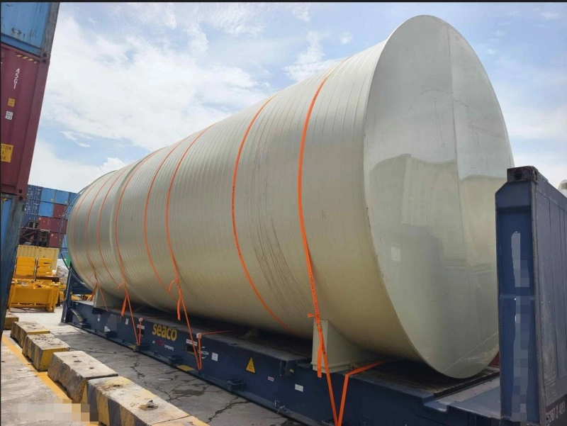 A massive beige cylindrical pressure vessel, wrapped in protective material and secured with bright orange tension straps, is mounted on a red flat rack container at a port yard, with stacked shipping containers visible in the background.