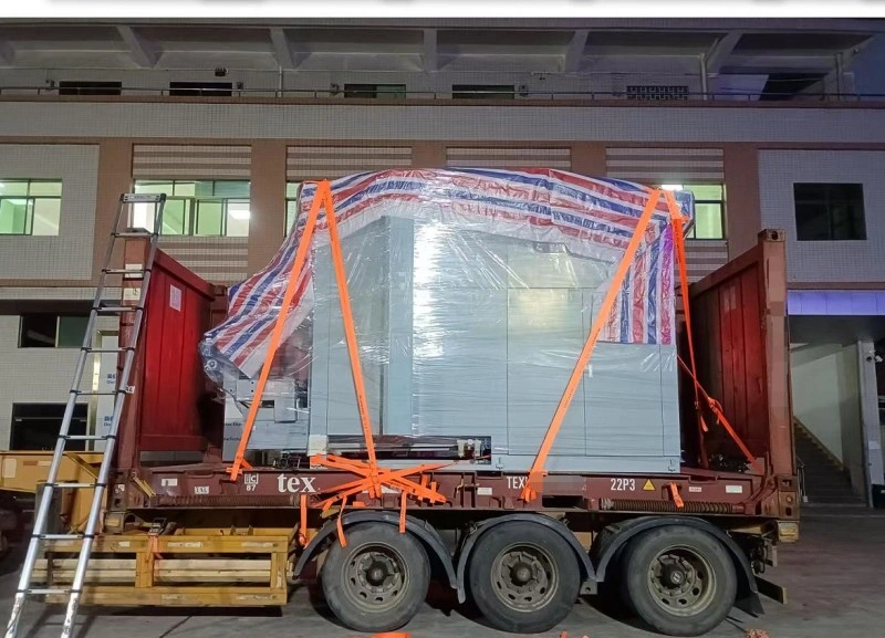 A massive gray electrical transformer unit, fully wrapped in clear plastic film with red-and-blue striped reinforcement tape, strapped down with bright orange webbing on a multi-axle flatbed trailer parked outside an industrial building at night.