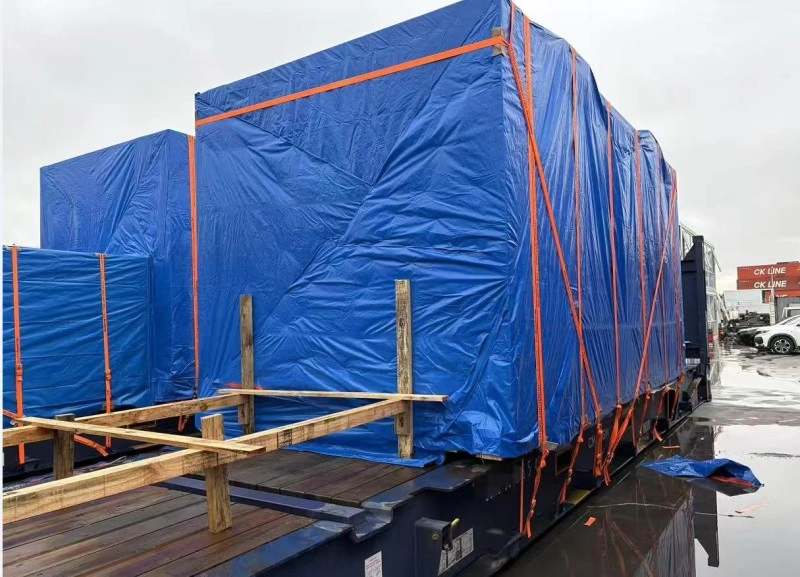 A massive box-shaped industrial unit, completely enveloped in a green tarpaulin secured with vertical orange straps and white tie-downs, sits atop a yellow flat rack container in an open yard; background shows stacked shipping containers and a yellow pickup truck under partly cloudy sky.