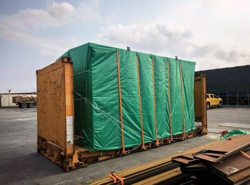 A massive box-shaped industrial unit, completely enveloped in a green tarpaulin secured with vertical orange straps and white tie-downs, sits atop a yellow flat rack container in an open-air depot under partly cloudy skies.