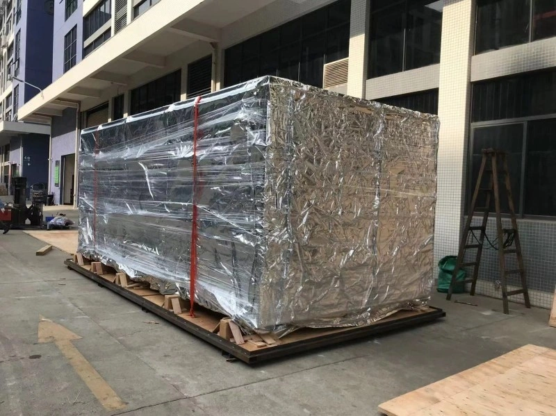 A white industrial machine, possibly a CNC machine or large pump, is loaded onto a grey flat rack container. The machine is secured with orange lashing straps, and the background shows a busy port yard with stacked shipping containers from companies like Maersk and Hamburg Sud.