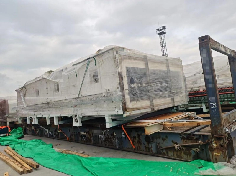 A massive white industrial machine, fully encased in transparent plastic wrap and secured with green straps, rests on a flat rack container at a port facility, ready for international transport under overcast skies.