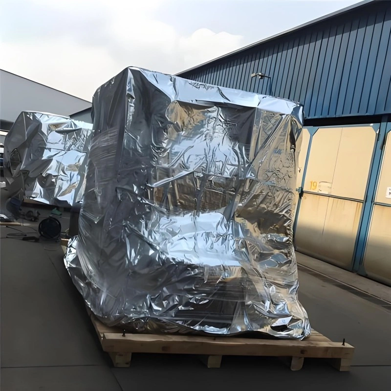 Heavy equipment wrapped in reflective aluminum foil packaging on a wooden pallet outside a factory, demonstrating moisture-proof protection against humidity and salt spray.