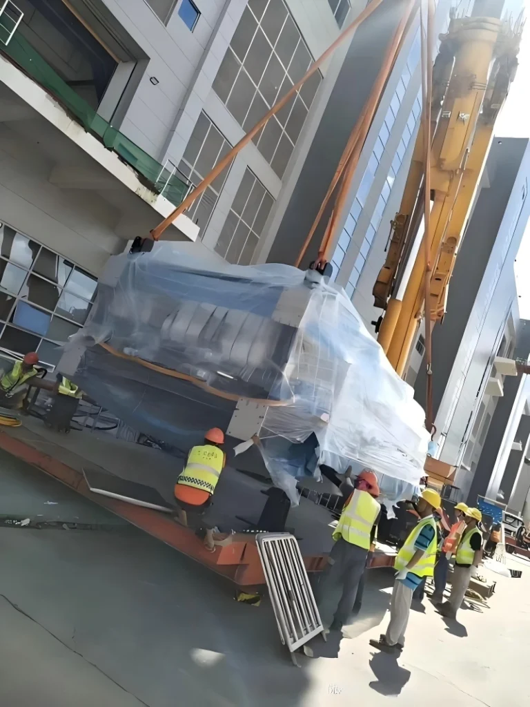 A mobile crane lifting a wrapped industrial machine while workers in safety vests coordinate the operation outside a modern factory building.