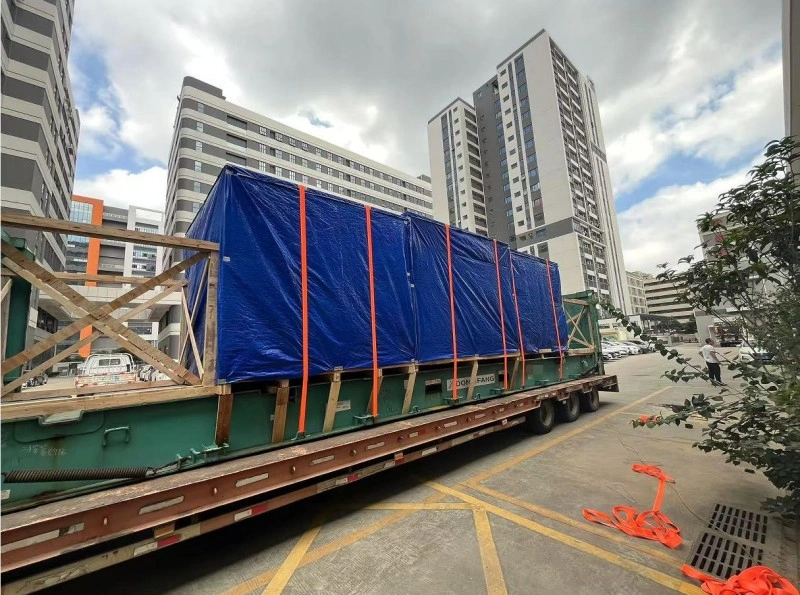 Side view of a lowboy trailer transporting a massive machine covered in blue tarpaulin and secured with orange straps, navigating through a modern urban area with tall buildings and scattered construction materials nearby.