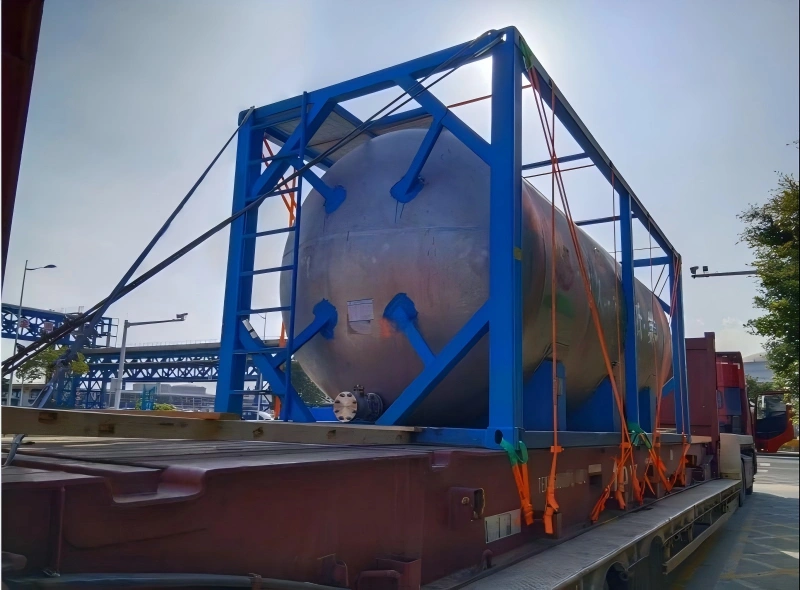 A large, cylindrical stainless steel pressure vessel is secured within a custom blue steel support frame on a flatbed trailer. The frame is lashed to the trailer with multiple orange ratchet straps, and the vessel features visible flanges and nozzles. The scene is set outdoors on a sunny day near a port or industrial area.