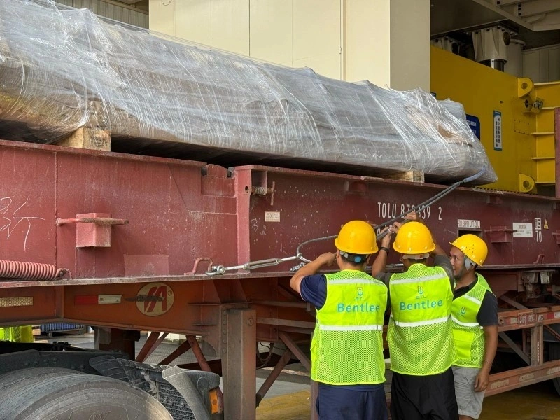 Three workers wearing yellow hard hats and green "Bentlee" safety vests are tightening steel wire ropes and turnbuckles on a red flatbed trailer carrying a large wrapped cylindrical load; close-up view of manual securing operations.
