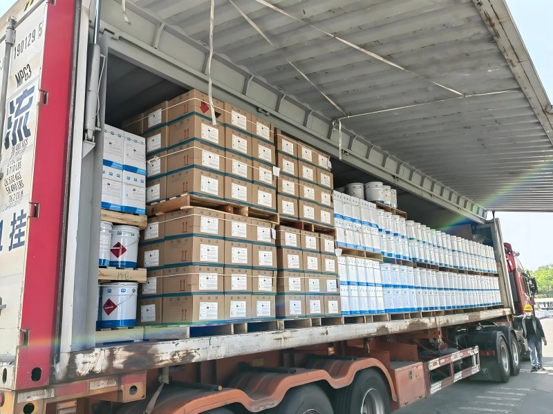 Stacked cardboard boxes and white metal drums with hazard labels loaded on pallets inside a semi-trailer truck