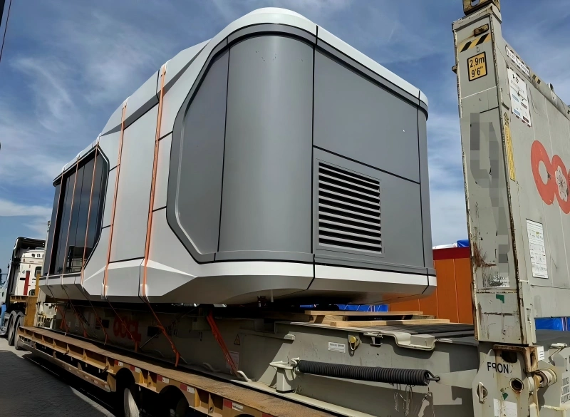 A sleek, grey and white prefabricated capsule house with large windows is secured with orange straps on a flatbed trailer, next to a shipping container in a transport yard