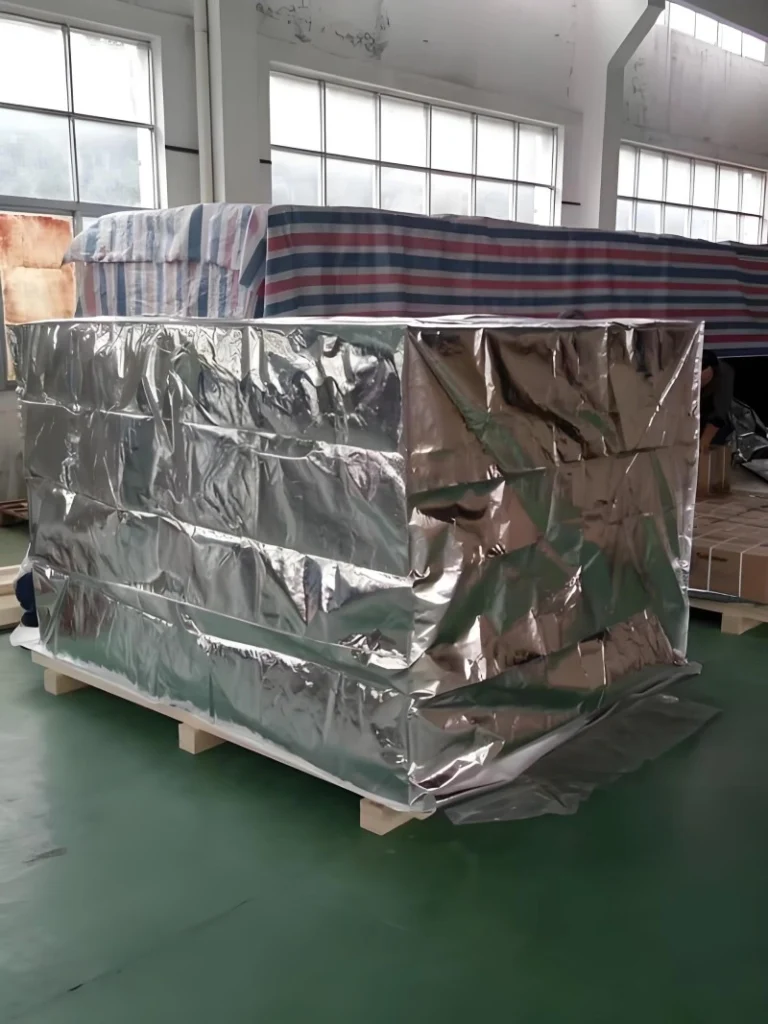 Equipment encased in shiny aluminum foil vacuum wrap with additional striped protective fabric covering above, mounted on a wooden pallet inside a factory setting, illustrating comprehensive export packaging strategy.
