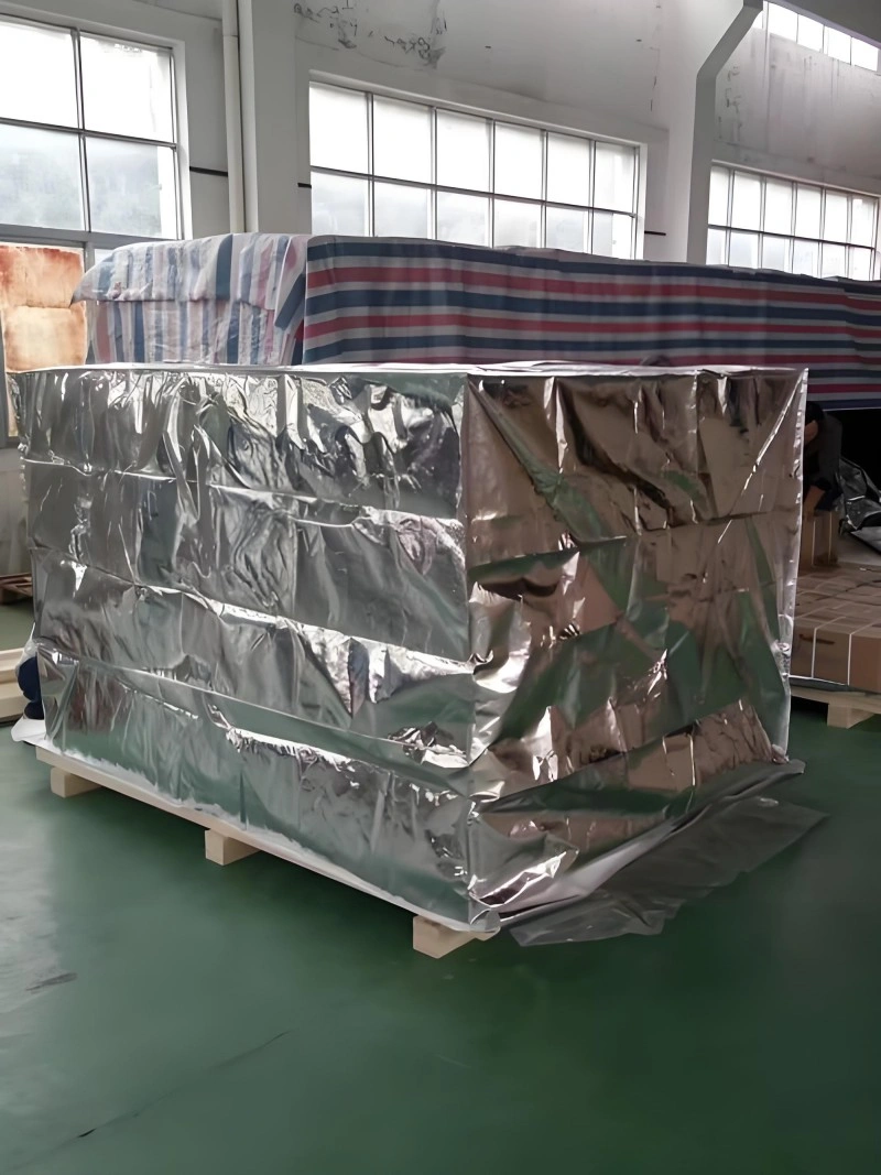 Equipment encased in shiny aluminum foil vacuum wrap with additional striped protective fabric covering above, mounted on a wooden pallet inside a factory setting, illustrating comprehensive export packaging strategy.