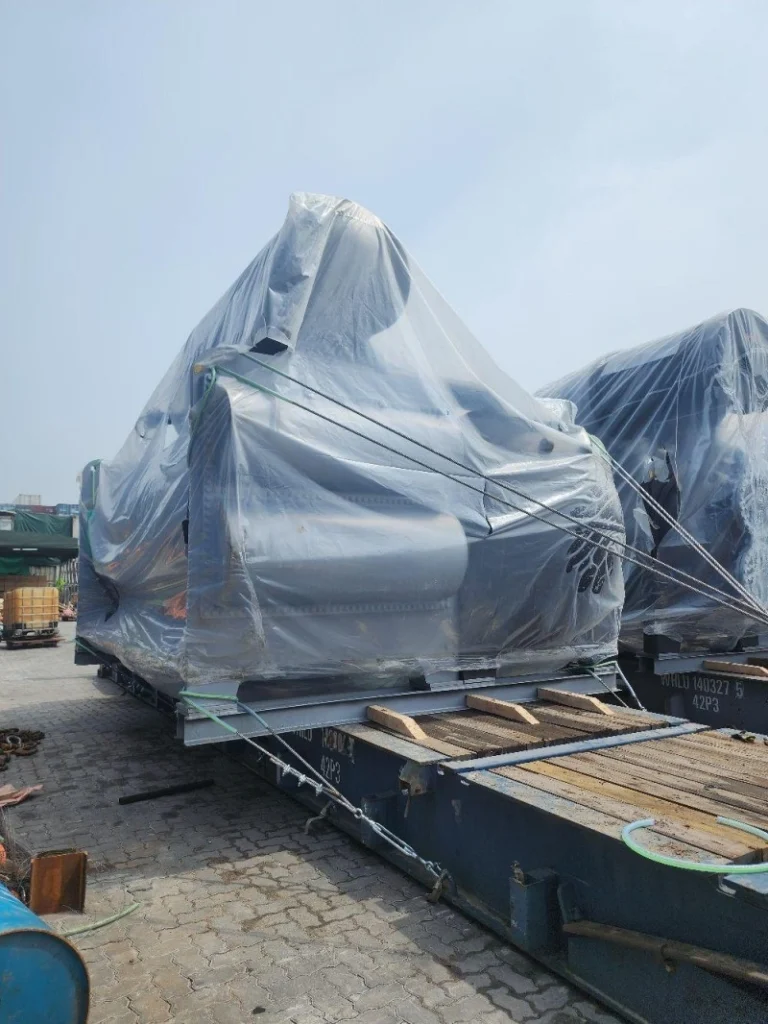Two large pieces of industrial machinery, each wrapped in clear protective plastic, are secured on separate blue flat rack containers in a shipping yard. The cargo is positioned with wooden dunnage and steel wire lashing.