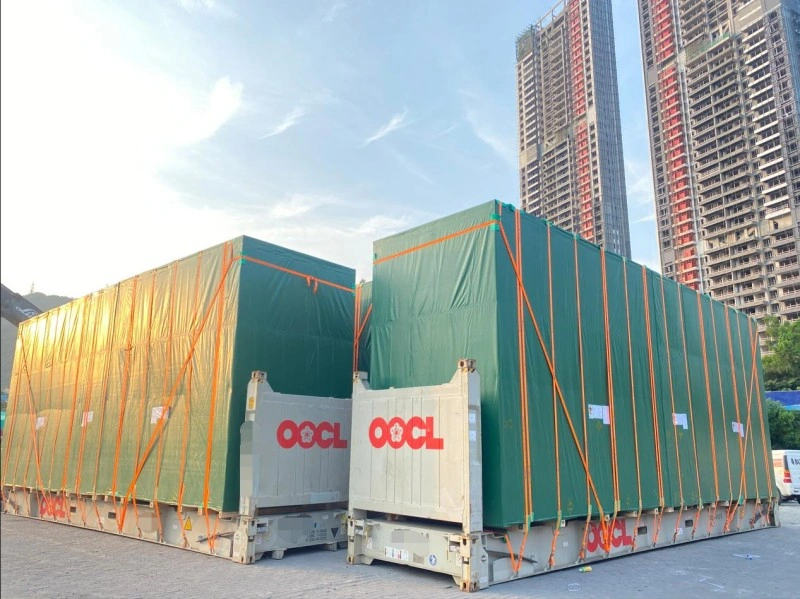 Two large OOCL-branded flat rack containers loaded with oversized industrial equipment covered in green protective tarps and secured with orange lashing straps, positioned in an urban logistics yard with high-rise buildings in the background.