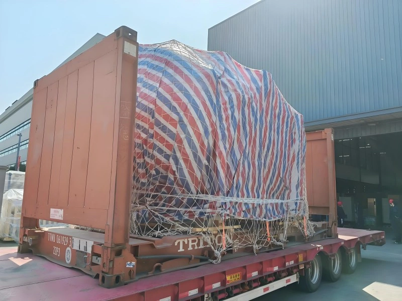 Heavy industrial equipment secured inside an open-top shipping container using a red, white, and blue striped tarpaulin and safety netting, illustrating specialized transport solutions for oversized or top-loaded cargo.