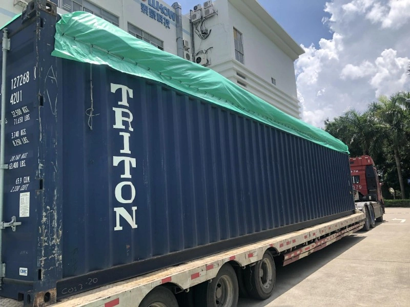 A blue open top shipping container covered with a green tarpaulin is mounted on a flatbed truck, parked near an industrial facility under bright daylight conditions.