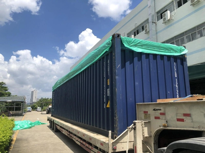 A blue open top shipping container covered with a green tarpaulin is being transported on a flatbed truck outside an industrial facility under a partly cloudy sky, indicating preparation for export or relocation.