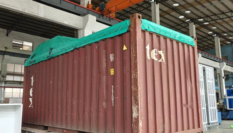 A red open top container with a green tarpaulin cover is positioned in a warehouse, ready for loading oversized industrial equipment. The container features visible structural reinforcements and safety markings.