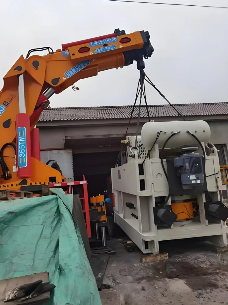 Workers in safety gear guiding a large wooden crate being lifted by a yellow crane onto a flatbed truck outside a modern factory building.