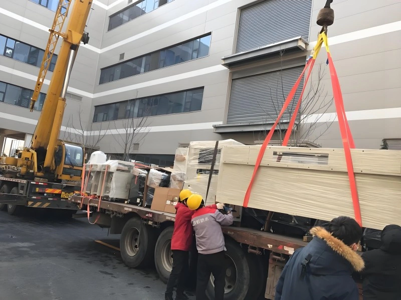 Workers in safety helmets guiding a large wooden crate suspended by orange lifting straps from a yellow crane outside a modern industrial building.