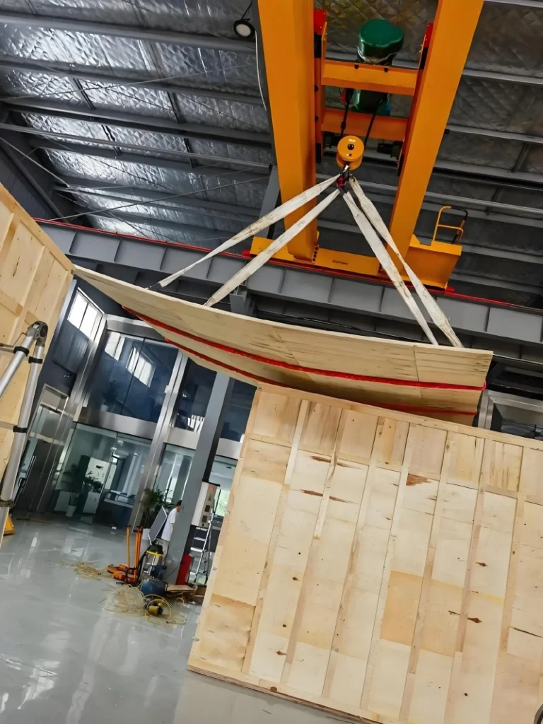 An orange gantry crane lifting a curved plywood lid into position over a partially assembled wooden export crate inside a modern factory, highlighting precision engineering in custom packaging construction.