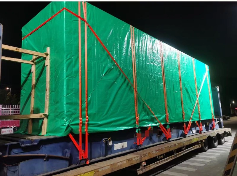 A large, rectangular cargo unit is covered with a green waterproof tarp and secured on a blue flatbed trailer using multiple red lashing straps and wooden support frames, shown at night under artificial lighting.