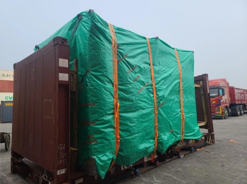 A large, box-shaped cargo unit, covered in a green waterproof tarp, is loaded onto a brown flat rack container. Multiple orange lashing straps are tightly secured around the cargo, with wooden support structures visible at the front and rear, and a red truck in the background.