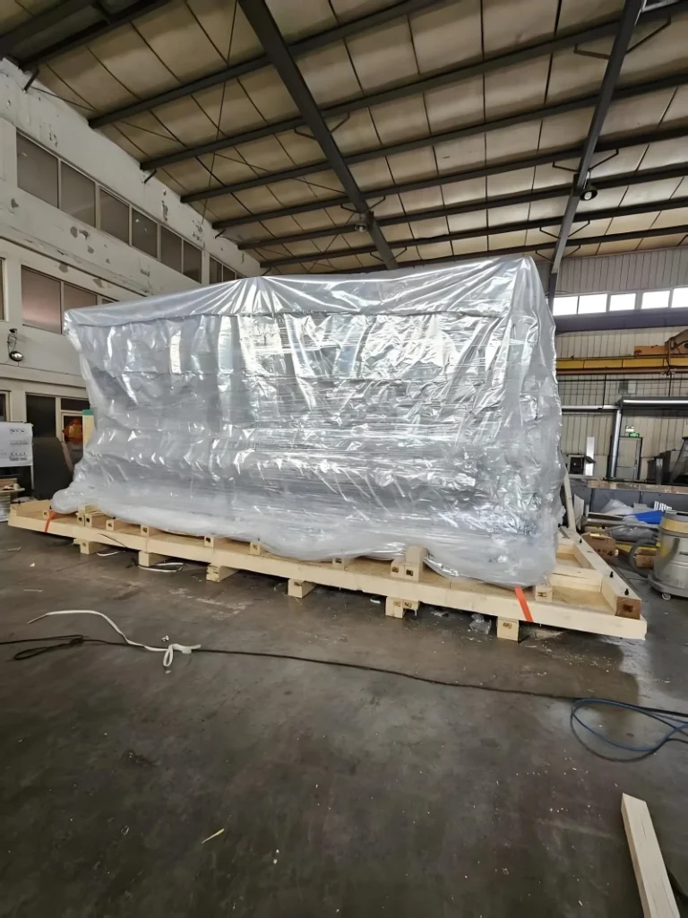 Massive machine covered in transparent plastic wrap on custom wooden skid, prepared for open-top container or flat rack loading with visible lifting points and structural support.