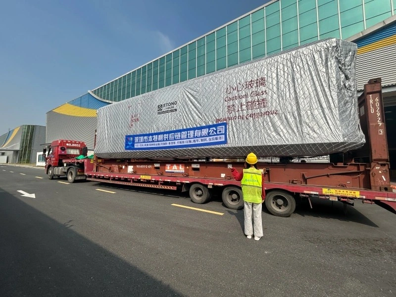 A massive glass panel wrapped in silver protective material and labeled “Caution: Glass” is being transported on a lowboy trailer outside a modern factory building under clear skies.