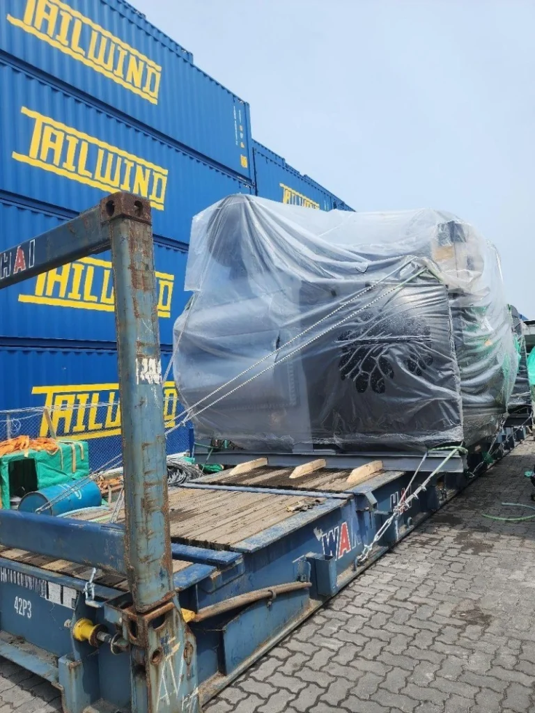 An irregularly shaped industrial machine, covered in protective film and anchored with diagonal steel cables, sits atop a blue flat rack container adjacent to stacked “TAILWIND” branded shipping containers in a busy port terminal.