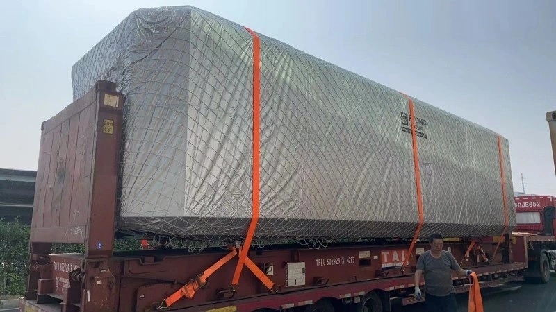 A massive rectangular cargo unit fully encased in silver reflective insulation wrap and secured with orange webbing straps, loaded onto a red skeletal trailer; a logistics worker stands nearby monitoring the load.
