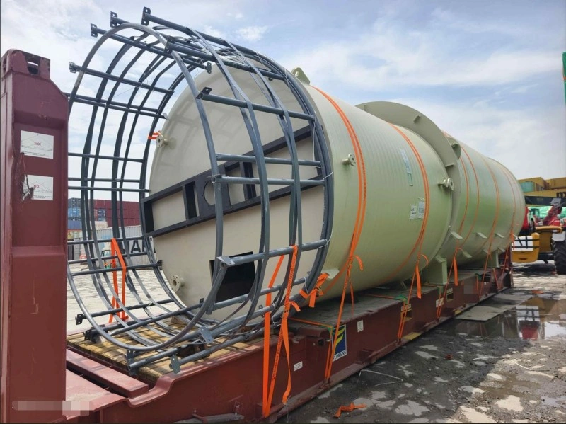 A long, light-green cylindrical pressure vessel, fitted with a robust steel cage frame at one end and bound by orange straps, is positioned on a red flat rack container in an outdoor logistics yard, with wet pavement indicating recent rain.