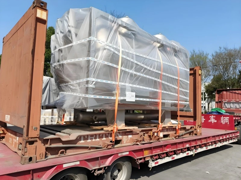 Side view of a massive industrial unit encased in protective plastic vacuum packaging and strapped securely onto a specialized flatbed trailer, positioned next to a shipping container frame for international transport.