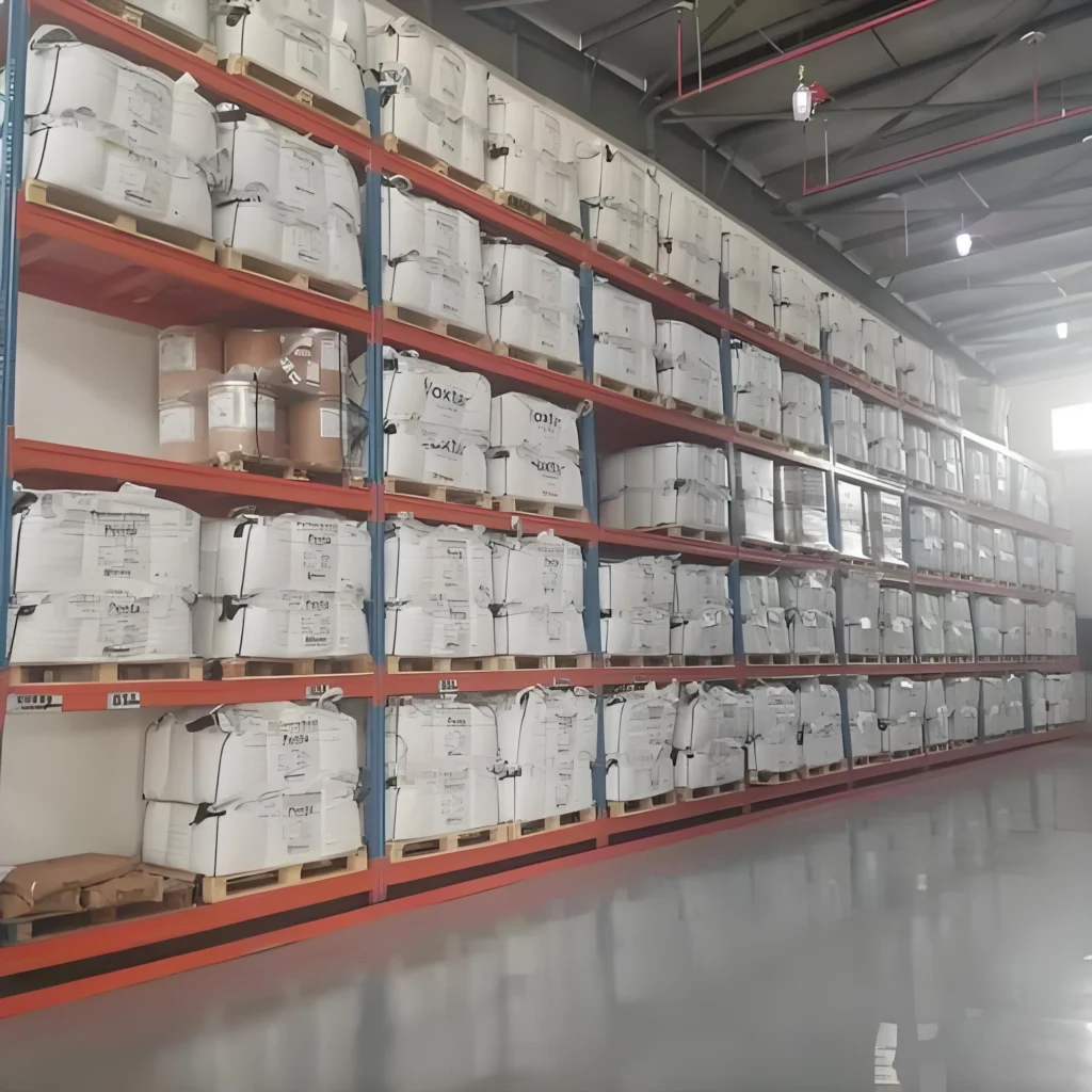 High-bay warehouse racking system storing numerous white bulk bags on wooden pallets, with some brown paper sacks visible. The organized storage setup is typical for safe handling of industrial materials in a logistics environment.