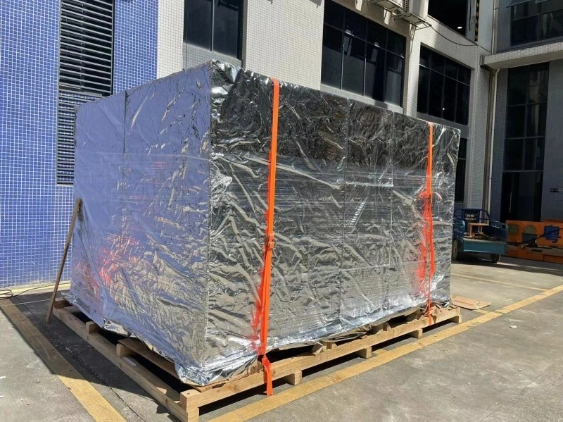 A large, rectangular cargo unit is placed on a wooden pallet and fully wrapped in a reflective silver protective film, secured with two bright orange lashing straps, positioned outdoors in front of a modern industrial building with blue-tiled walls.