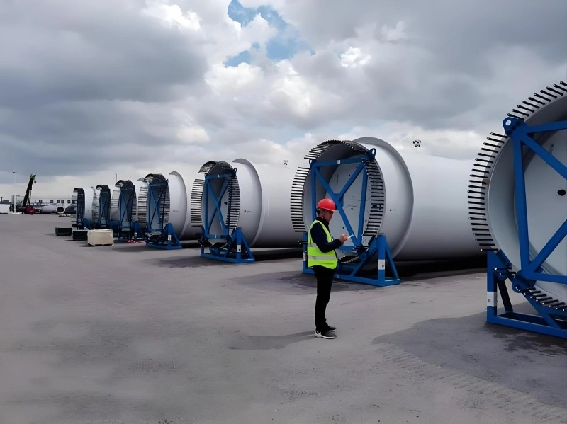 A worker in high-visibility vest and hard hat inspects large cylindrical wind turbine tower sections mounted on blue support frames in an open logistics yard under cloudy skies, highlighting quality control before international shipment.