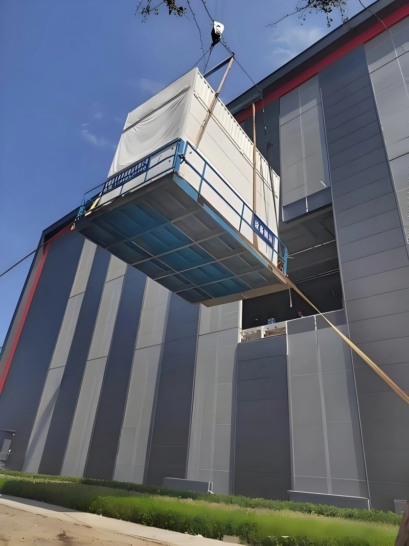 Workers in safety helmets supervising the crane lifting of a large industrial machine inside a factory workshop, with other equipment visible in the background.