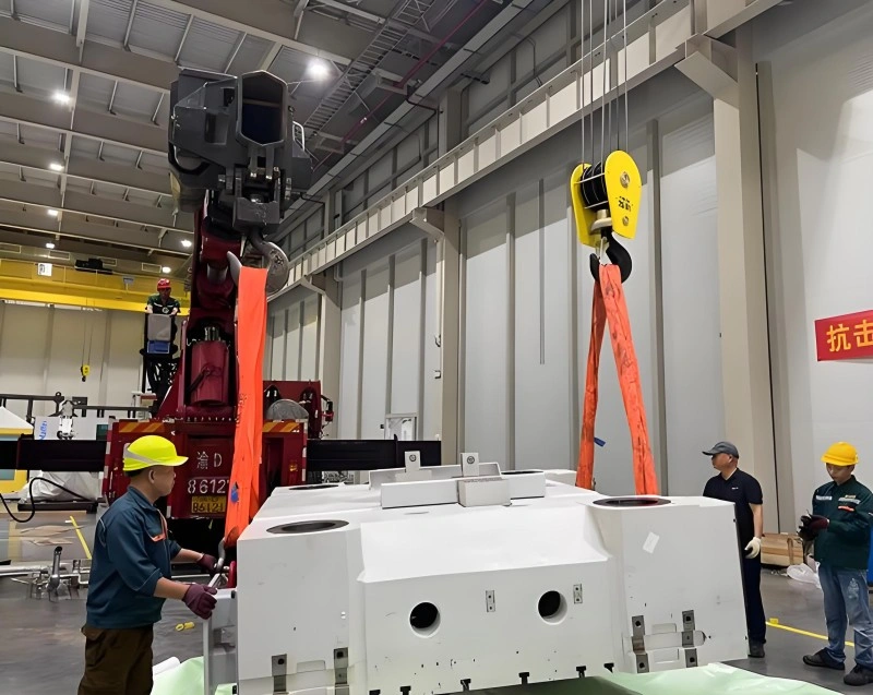 Workers guiding a large white machine component into position using an overhead crane inside a well-lit industrial facility.
