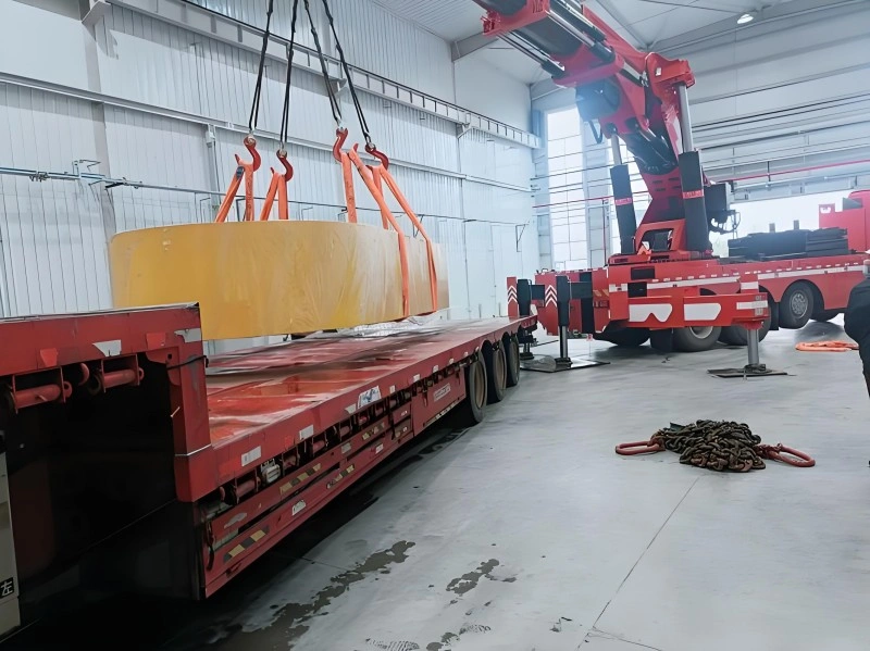 A red mobile crane lifting a large piece of industrial equipment inside a factory warehouse, with workers guiding the load safely onto a flatbed truck.
