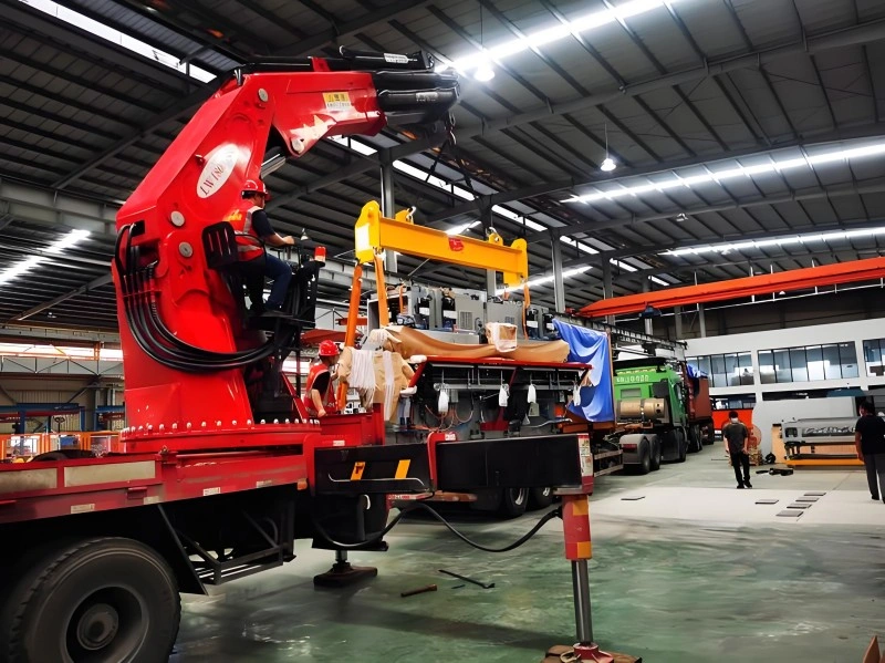 A red mobile crane lifting a large piece of industrial equipment inside a factory warehouse, with workers guiding the load safely onto a flatbed truck.
