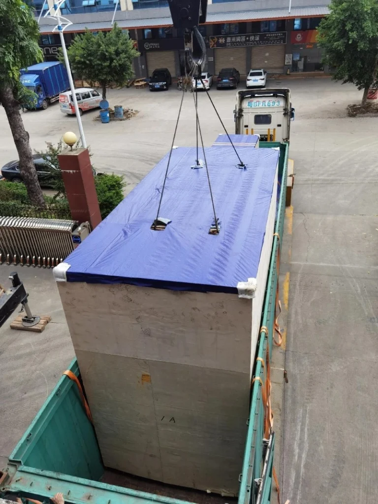 Crane lowering a large wooden crate covered with blue tarp into an open-top shipping container, demonstrating careful coordination for OOG freight.
