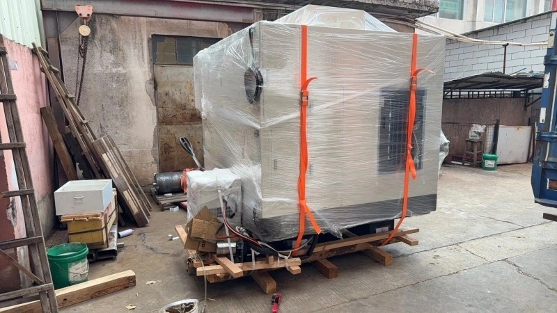 A large industrial CNC machine wrapped in clear plastic film and secured with orange straps on a wooden pallet, ready for transport.