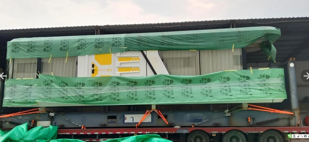 A side view of a white and yellow prefabricated capsule house, partially covered with green protective film, secured with orange straps on a flatbed trailer in a warehouse setting